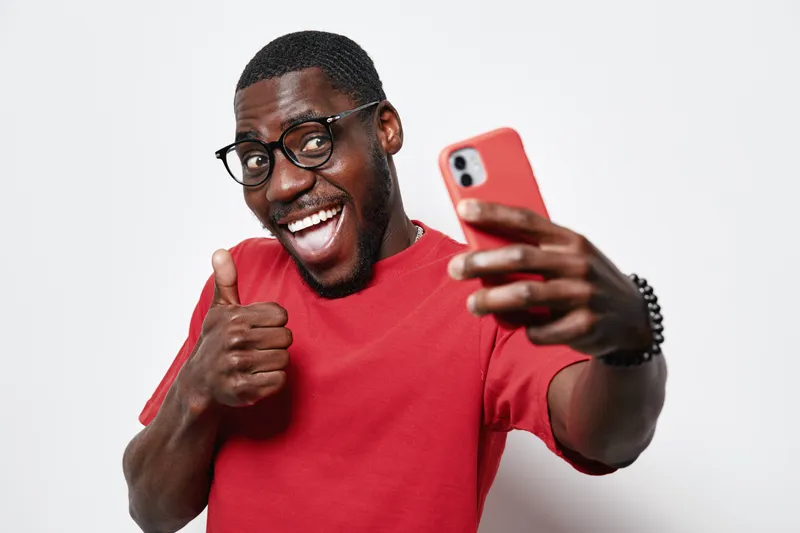 African American man with dark skin wearing glasses and red shirt taking selfie with smartphone showing thumbs up gesture and smiling on white background.