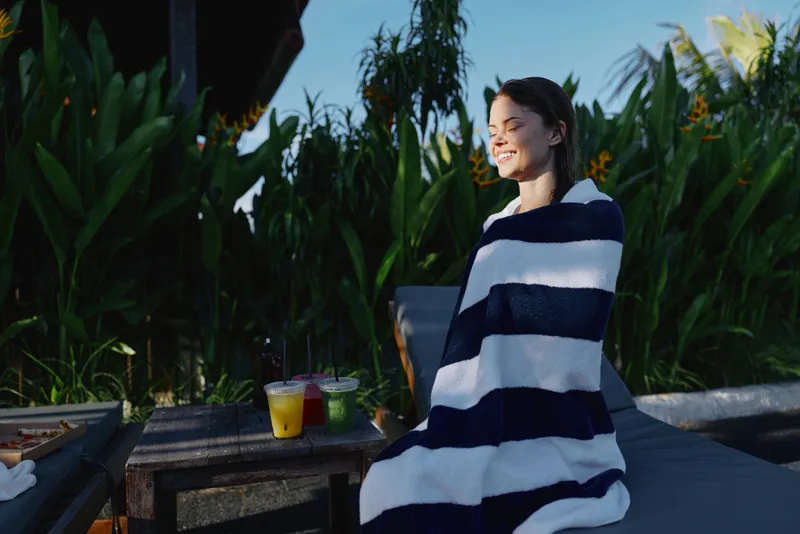 Cheerful woman outdoors, wrapped in a striped towel, enjoying sunlit relaxation, lush greenery backdrop, casual leisure moment, soft breeze, natural colors and warm lighting, soft textures