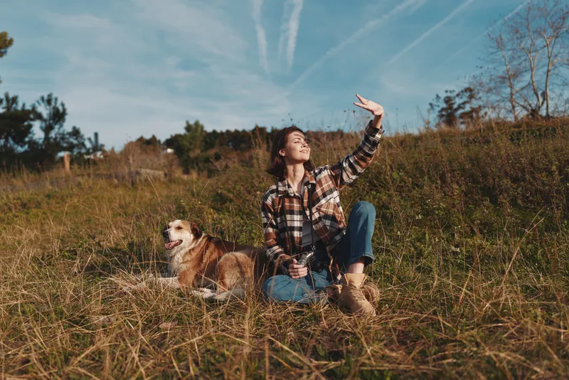 Woman and dog in an open field under a blue sky, enjoying nature and sunshine. Casual plaid shirt, jeans and boots as she sits and waves toward the bright horizon.