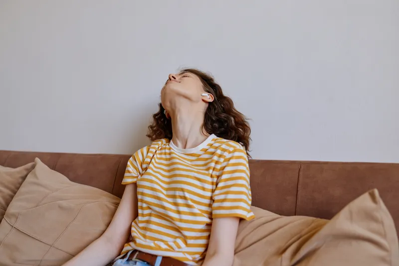 Young woman in a yellow striped t shirt sits on a sofa, eyes closed, head tilted back, enjoying a relaxed, carefree moment indoors with warm natural light.