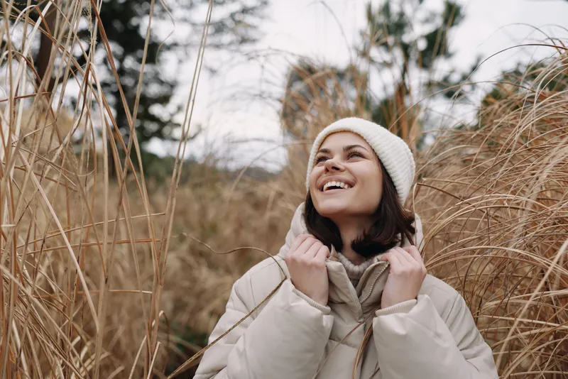 Woman in a warm beige coat and knit beanie stands among tall grasses, smiling and looking up, capturing a cozy outdoor winter moment in natural light.