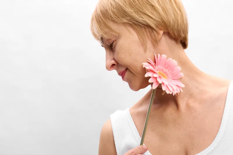 Woman blonde studio portrait, holding pink flower petals to her neck, side profile smile, gentle expression, white background, soft light, minimal composition, closeup skincare