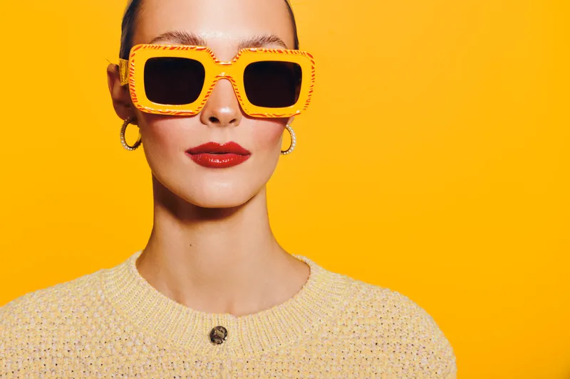 Stylish woman with orange sunglasses and red lipstick poses against bright yellow background. Fashion concept with vibrant colors and confident expression in studio lighting.