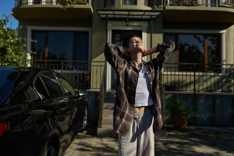 A young woman stands outside a modern building, arms raised to the sky, wearing a plaid shirt over a white tank and light trousers, enjoying a carefree moment in bright sunlight