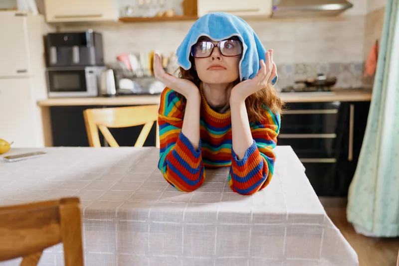A bored woman sits at a dining table in a bright kitchen, wearing a striped sweater and a towel on her head, eyes upward, resting chin on hands, capturing a playful home moment and everyday routine.