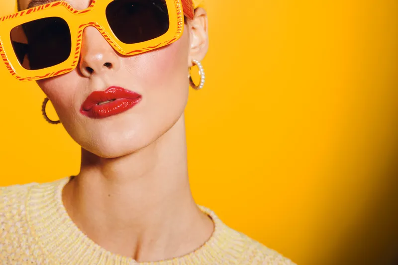 Stylish woman with bright orange sunglasses and red lipstick poses against vibrant yellow background. Fashion portrait with trendy accessories and bold makeup for summer style concept.