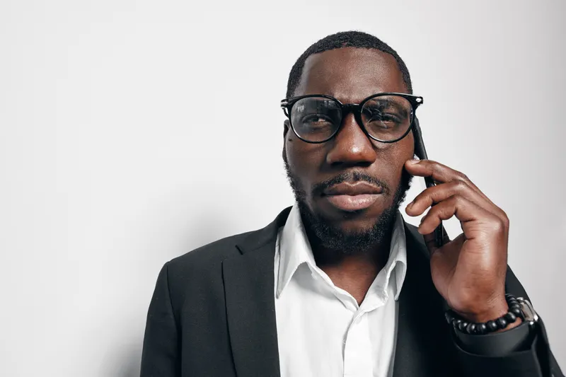 Black man with dark skin wearing glasses and suit talking on smartphone in office. Professional African American businessman with beard and white shirt using mobile phone for communication.