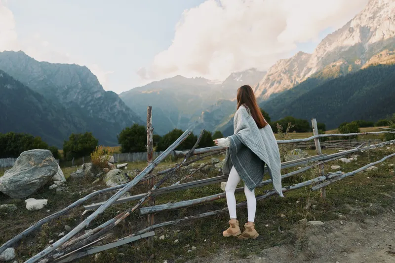 Young woman wrapped in a cozy blanket stands near a wooden fence looking at distant mountains under a cloudy sky during a cool day.