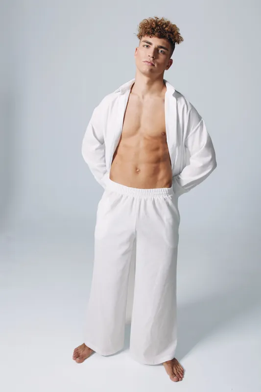 Young shirtless man with curly hair wearing loose white pants and unbuttoned white shirt posing standing barefoot on grey background with confident expression and hands behind back.