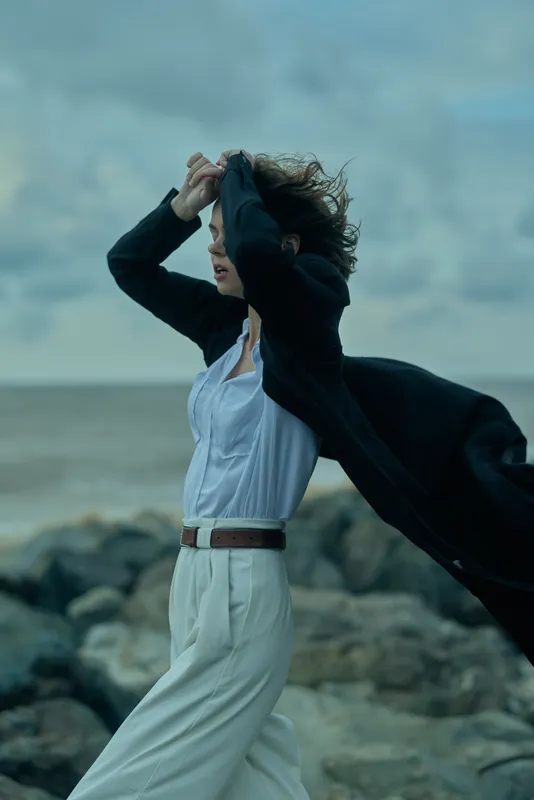 Windblown businesswear along the seaside, a flowing white suit with a navy coat billowing behind, rocky shoreline and waves framing a dynamic stride, conveying confidence, motion, and outdoor