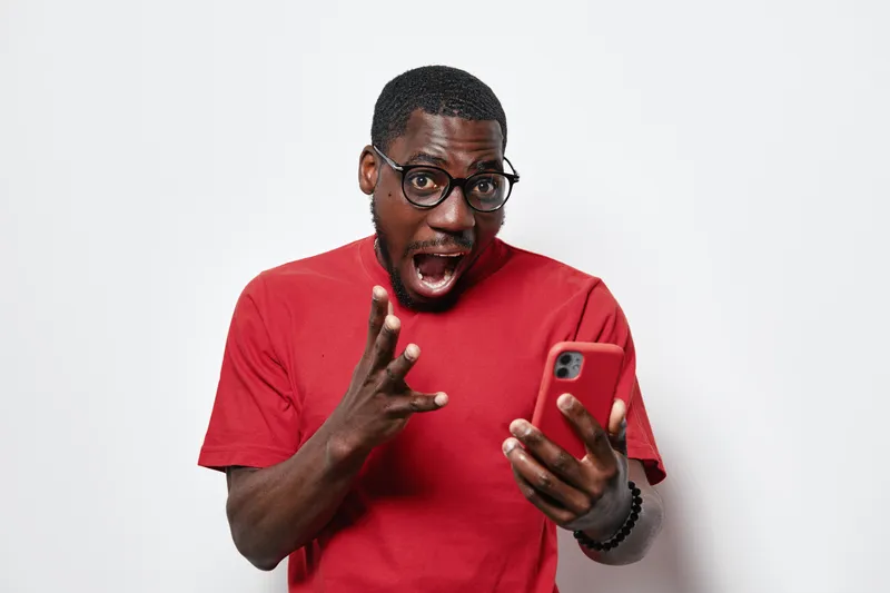 African American man with dark skin and glasses wearing red shirt looking surprised while holding smartphone in hand against white background. Casual technology and emotion.