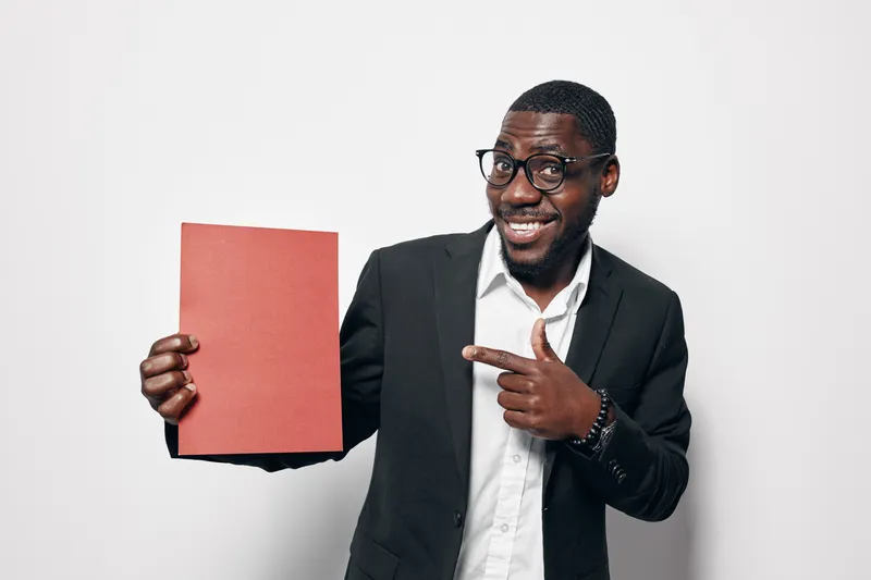 African American man with dark skin wearing glasses and suit holding red paper and smiling while pointing at it on white background. Business, presentation and confident.