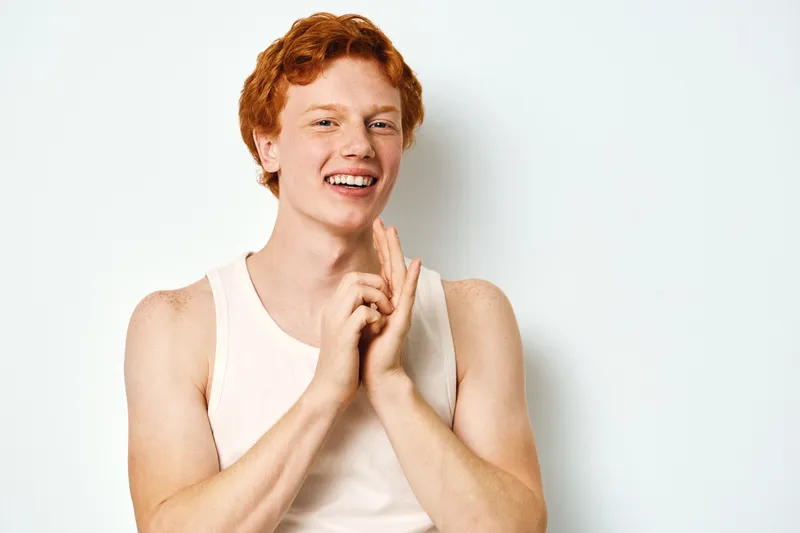 Redhead man with fair skin and freckles smiling and clapping hands wearing white tank top in studio with plain white background and casual appearance.