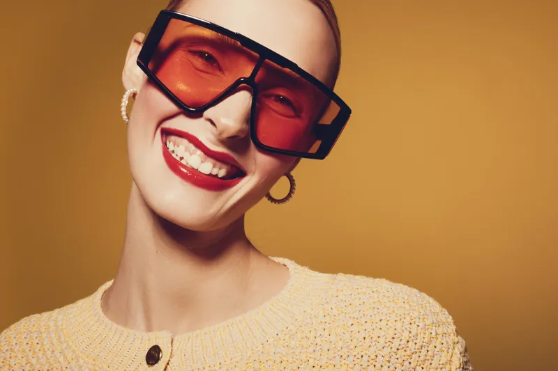 Smiling woman with red lipstick wears large orange sunglasses and yellow sweater. Close-up portrait on warm brown background. Concept of fashion, style, and confident beauty.