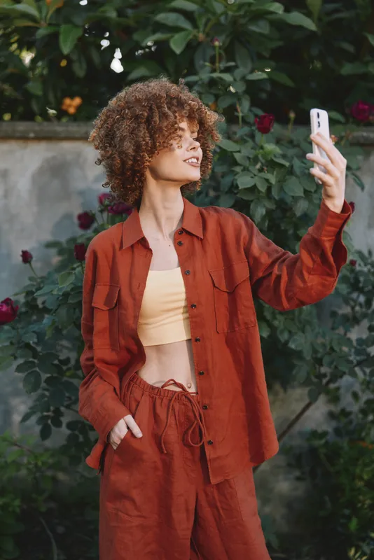 A stylish woman takes a confident selfie in an outdoor garden setting, wearing a rust colored matching outfit. The casual look and bright backdrop convey modern fashion and lifestyle vibes