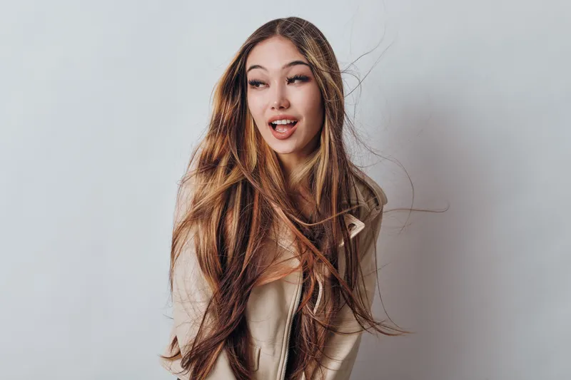 Young woman with long hair blowing in the wind wearing casual jacket and looking to side with playful expression on light background. Female model portrait with natural makeup and hairstyle.