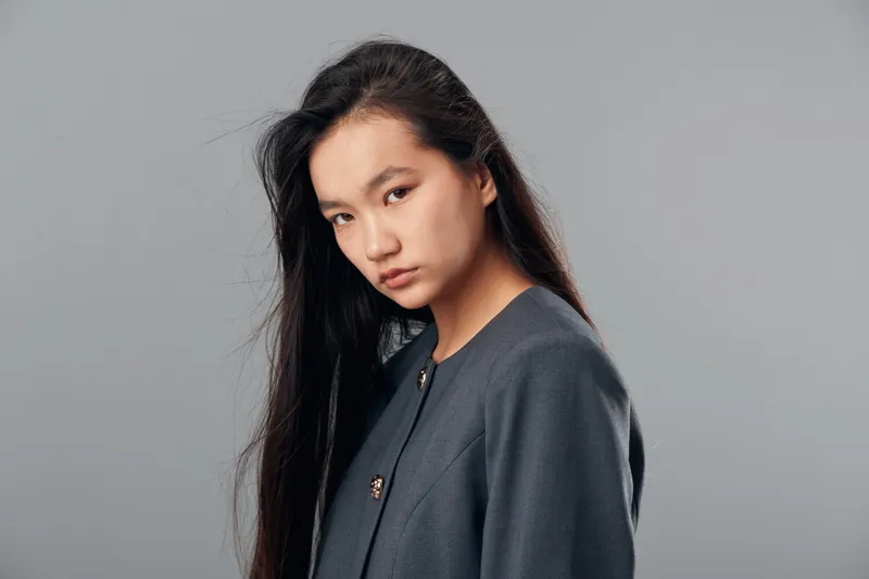 Young woman with long dark hair wearing a gray jacket posing against gray background in studio. Female model looking at camera with serious expression and natural makeup closeup portrait.