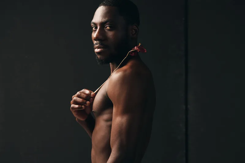 Creative conceptual portrait of African man with dark skin holding flower on shoulder in visual artistic style against black background. Male model with serious expression and bare torso.