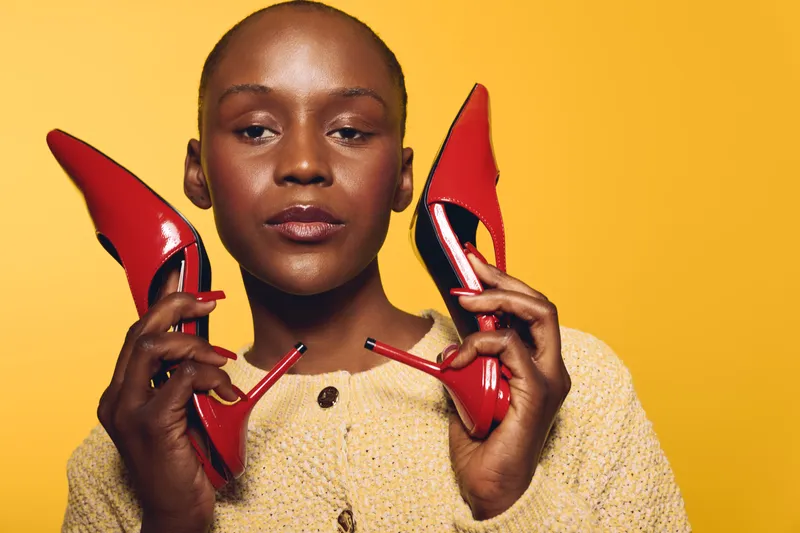 Portrait of confident black woman with dark skin holding red high heels near face against yellow background. Concept of fashion, style, beauty, and empowerment for dark skinned models.