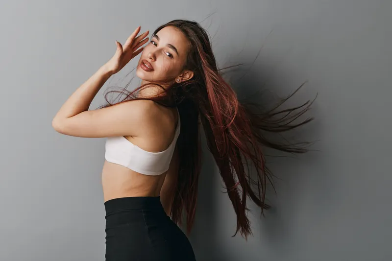 Woman with long hair posing in sportswear against gray background, showing movement and style. Female model in white top and black pants with natural makeup and confident expression.