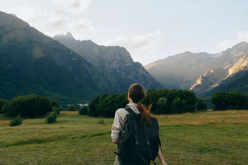 Young woman with a backpack gazes at majestic mountains over a green valley during a hike. Outdoor lifestyle, nature, exploration, peaceful mood.