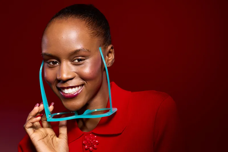 Smiling African woman with dark skin holding blue glasses near face wearing red outfit. Portrait on red background. Concept of fashion, beauty, confidence, and style for dark skin tone.
