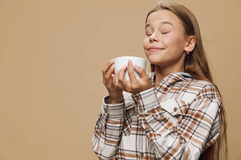 Girl with long hair in plaid shirt holding white cup with closed eyes and relaxed face enjoying hot drink against beige background with cozy atmosphere.