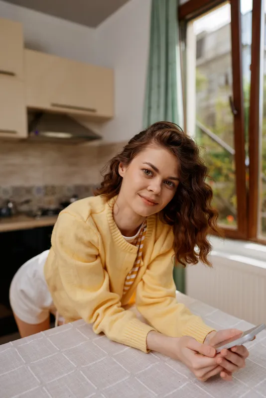 Young woman in a yellow sweater leans on a home kitchen table, smiling softly while using a smartphone, capturing a relaxed lifestyle moment in a bright, cozy indoor setting.