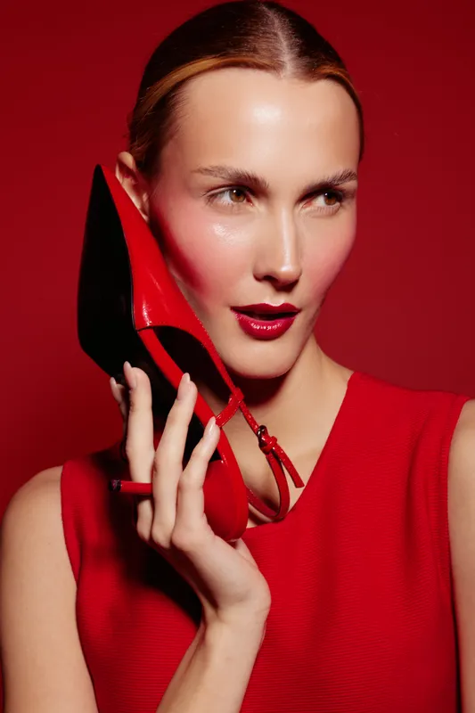 Stylish woman in red dress holds red high heel shoe near face on red background. Fashion concept with bold makeup and elegant accessories for modern glamour and style.