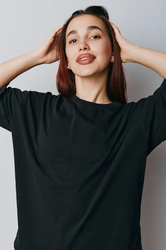 Young woman posing with hands behind head wearing black oversized shirt against neutral background. Casual style, confident expression and natural makeup for lifestyle portrait.