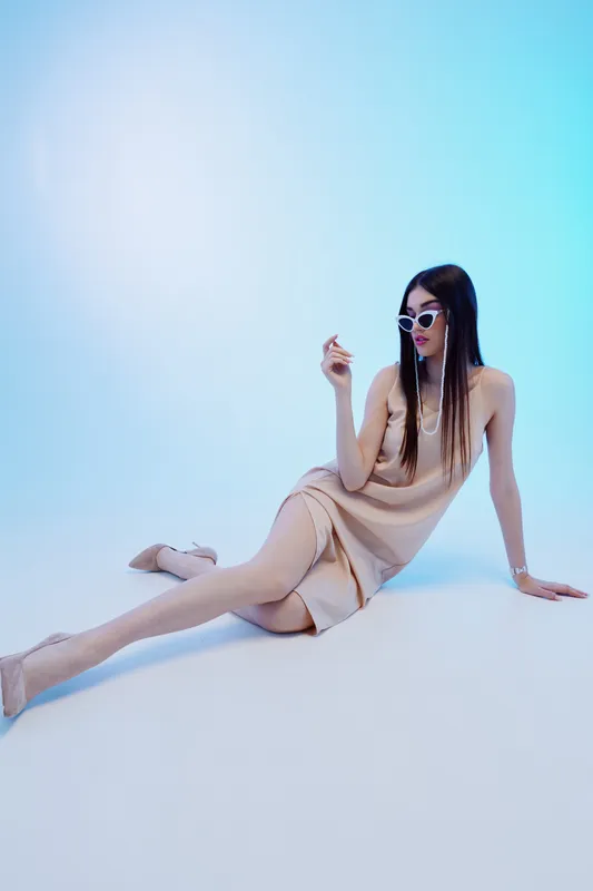 Fashionable woman in beige dress and sunglasses poses sitting on floor against blue background. Concept of beauty, fashion, style, and elegance in studio lighting.
