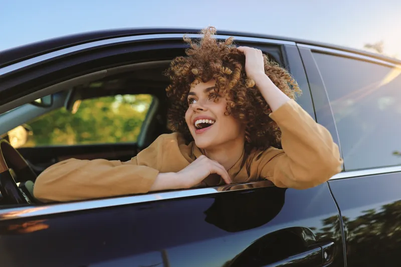 A stylish woman with curly hair leans from a car window, laughing softly in warm retro lighting. The scene conveys a carefree urban lifestyle with vintage color tones.