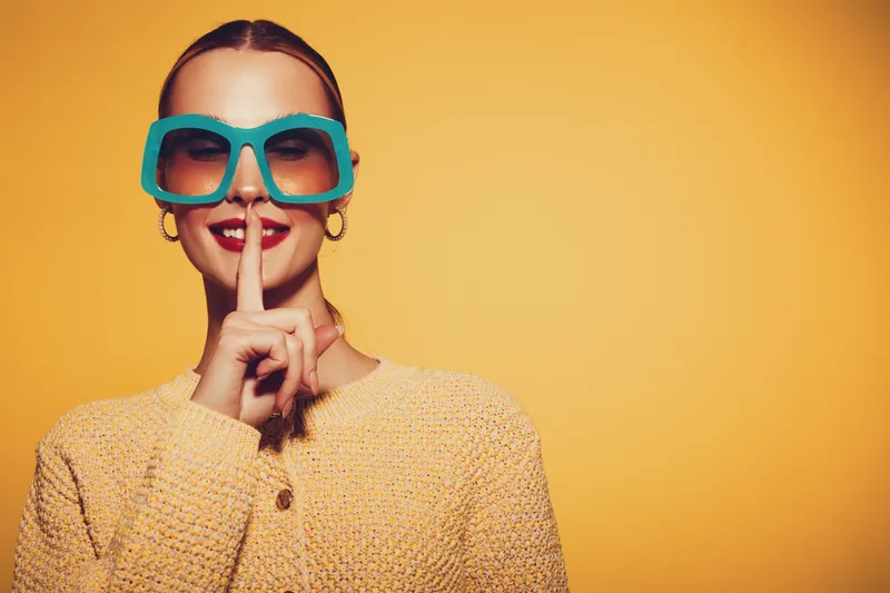 Young woman in oversized blue sunglasses and yellow sweater makes silence gesture with finger on lips against yellow background. Concept of secrecy, quiet, and playful mood.