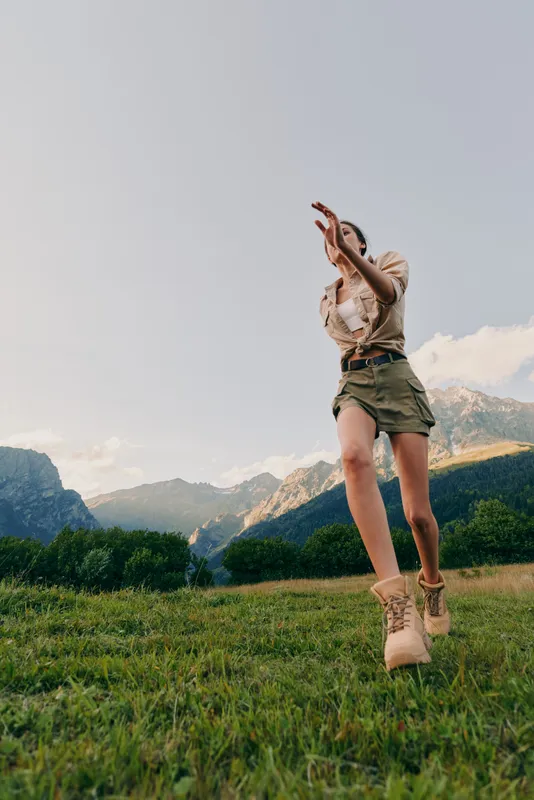 A young woman in casual clothes runs through a green meadow with mountains in the background under a clear sky. Concept of active lifestyle and freedom outdoors.