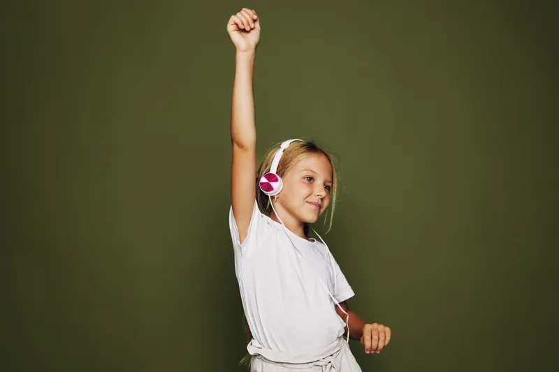 Young girl with headphones raising her arm and smiling. Studio shot on green background. Concept for music, joy, and childhood fun.