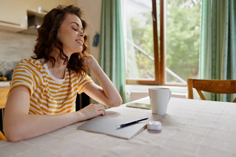 A middleaged woman sits at a home table with notebook, pen, and mug as sunlight streams through window and curtains, creating a calm, nostalgic moment