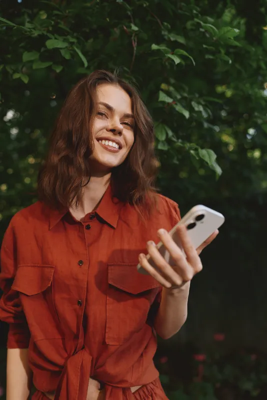 Cheerful woman outdoors, smiling while looking at her phone, sunlit scene, casual posture, natural colors, lush greenery background, candid lifestyle moment