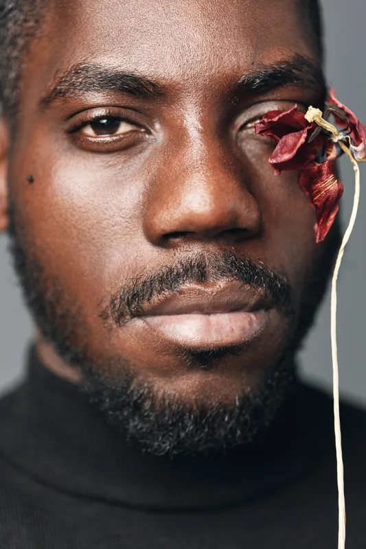 African man portrait closeup with dark skin and beard holding dried red flower near eye in creative conceptual visual style with serious expression and black turtleneck clothing.