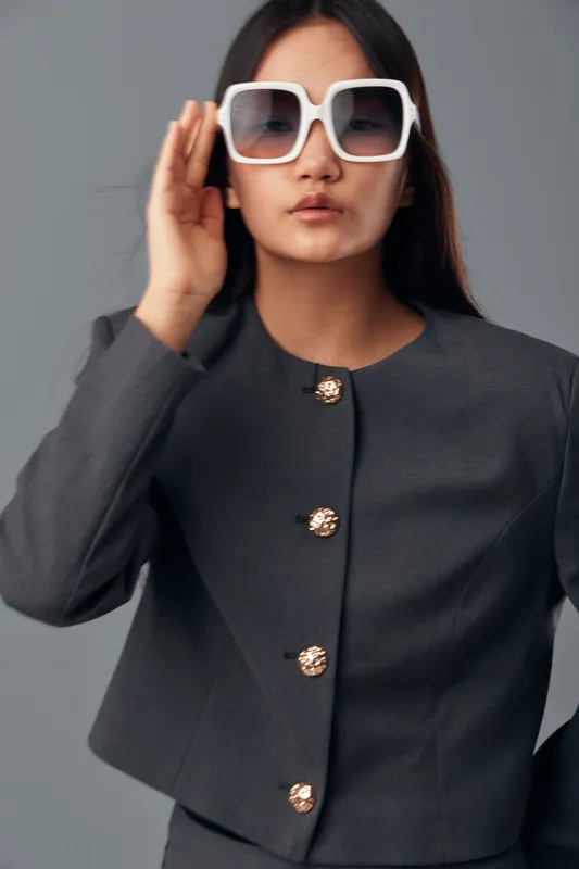 Woman with long dark hair wearing large white sunglasses and grey formal jacket with decorative buttons poses against grey background in studio. Fashion style portrait with confident expression.