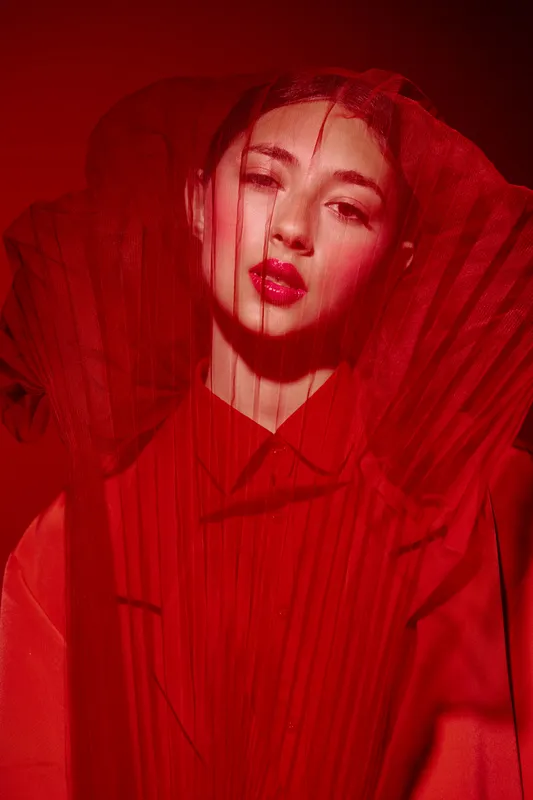 Fashion creative close up of woman with red lips wearing avantgarde high-neck dress and net veil on face in red hue studio light. Beauty portrait with dramatic and intense mood.