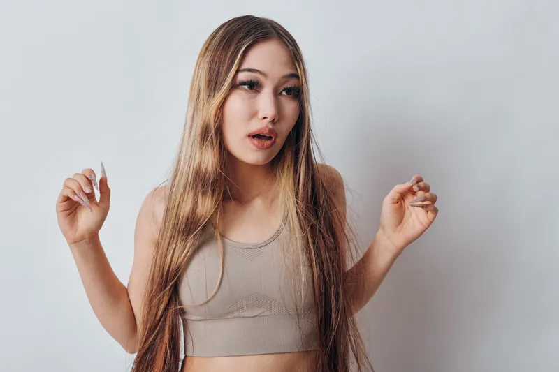 Young woman with long hair and braids wearing sleeveless top posing against white background with thoughtful expression and relaxed hands up.