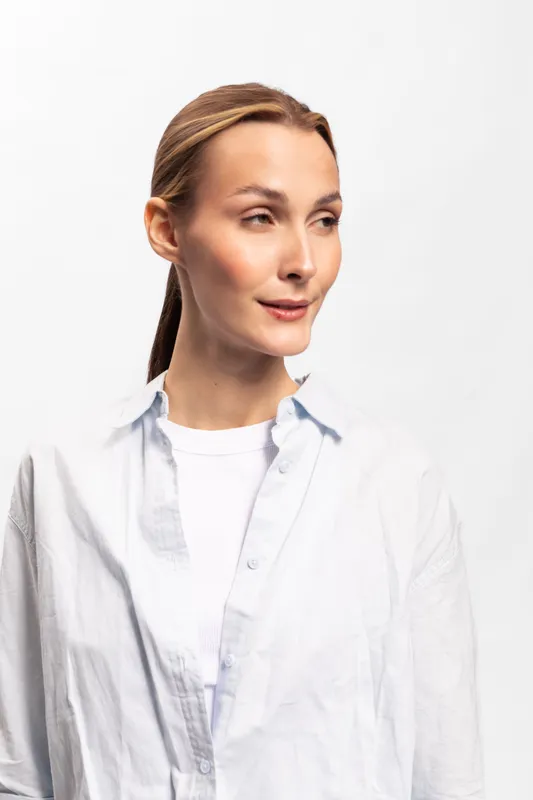Young woman with tied hair wears white shirt and t-shirt looks aside with calm expression on white background. Concept of casual style, natural beauty, and relaxed mood portrait.