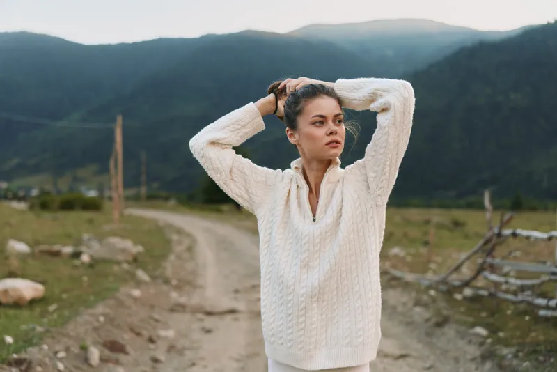 A woman in a white knitted sweater stands on a dirt path in a valley, hands in her hair, gazing into the distance with a calm, contemplative expression.