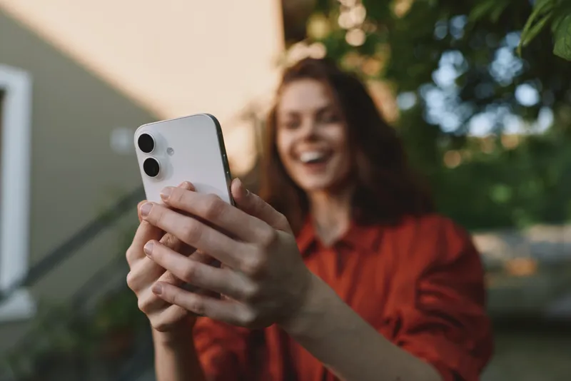 A smiling woman in casual wear enjoys a sunny outdoor moment, holding a phone for a selfie. A lifestyle scene about connection, joy, and modern daily routines