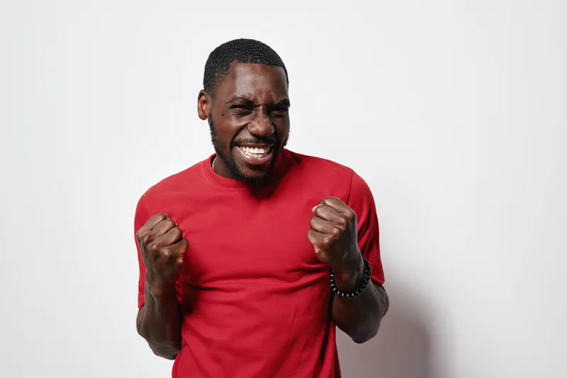African American man with dark skin wearing red shirt celebrating success with clenched fists and joyful expression on white background. Happiness and victory concept.