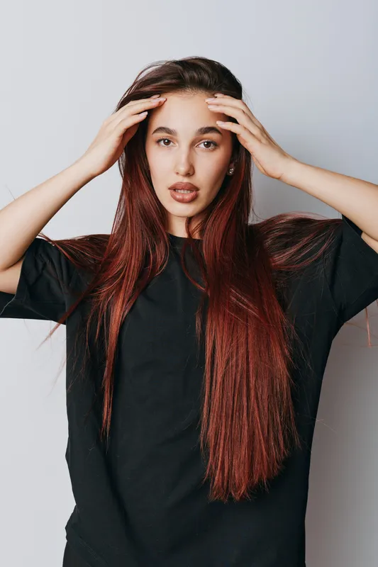 Young woman with long red hair wearing black shirt touching head with hands posing on white background. Portrait, emotion, casual style and beauty with natural expression and hairstyle.