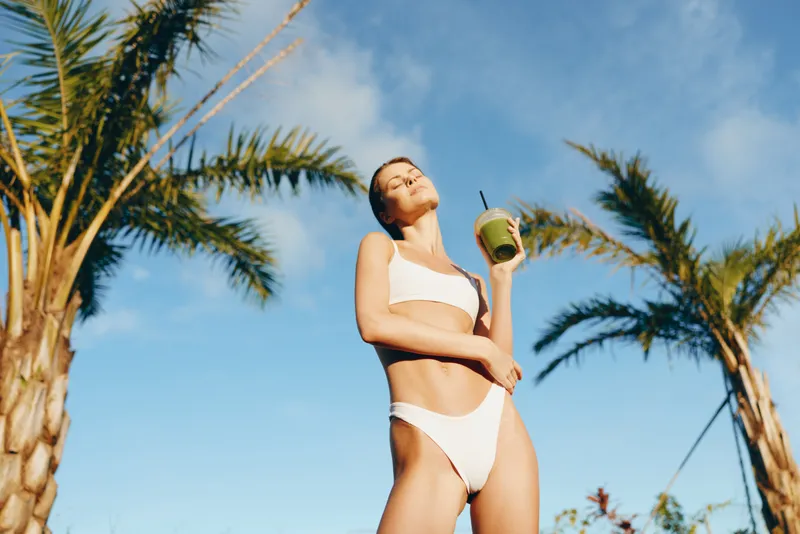 Young woman in a white bikini enjoys a green smoothie outdoors under palm trees on a sunny day with clear blue sky, expressing relaxation and wellness.