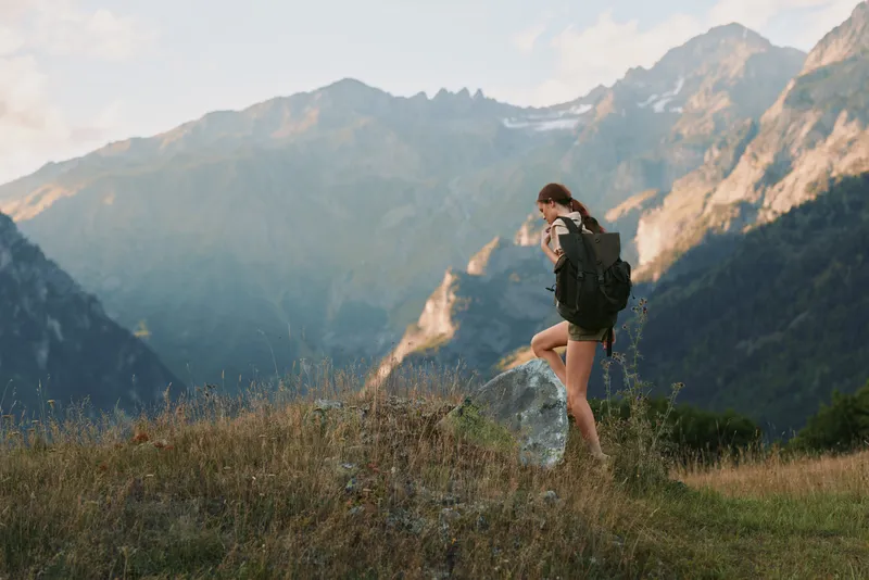 Young woman with a backpack hikes through a mountain meadow during a sunny day, enjoying nature and the scenic alpine landscape around her.