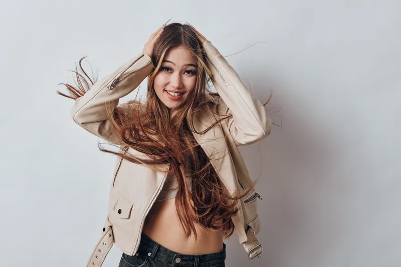 Young woman with long hair smiles joyfully while posing with hands in hair wearing beige jacket and black pants, isolated on gray background in studio.