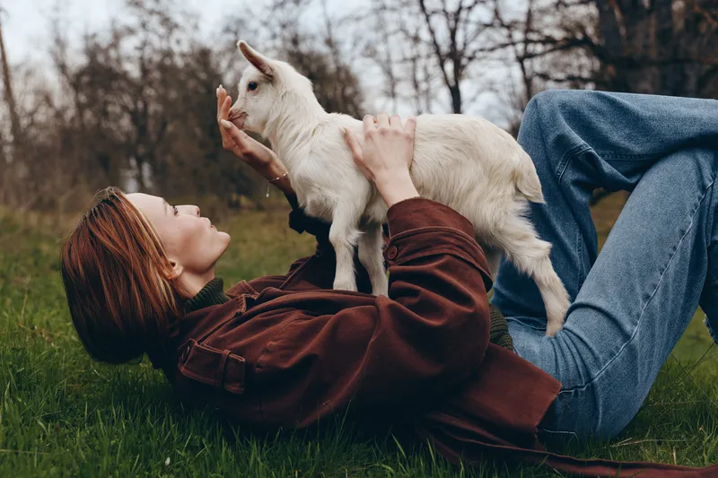Young woman lying on grass plays joyfully with a white baby goat in an outdoor natural setting during a relaxed lifestyle moment.